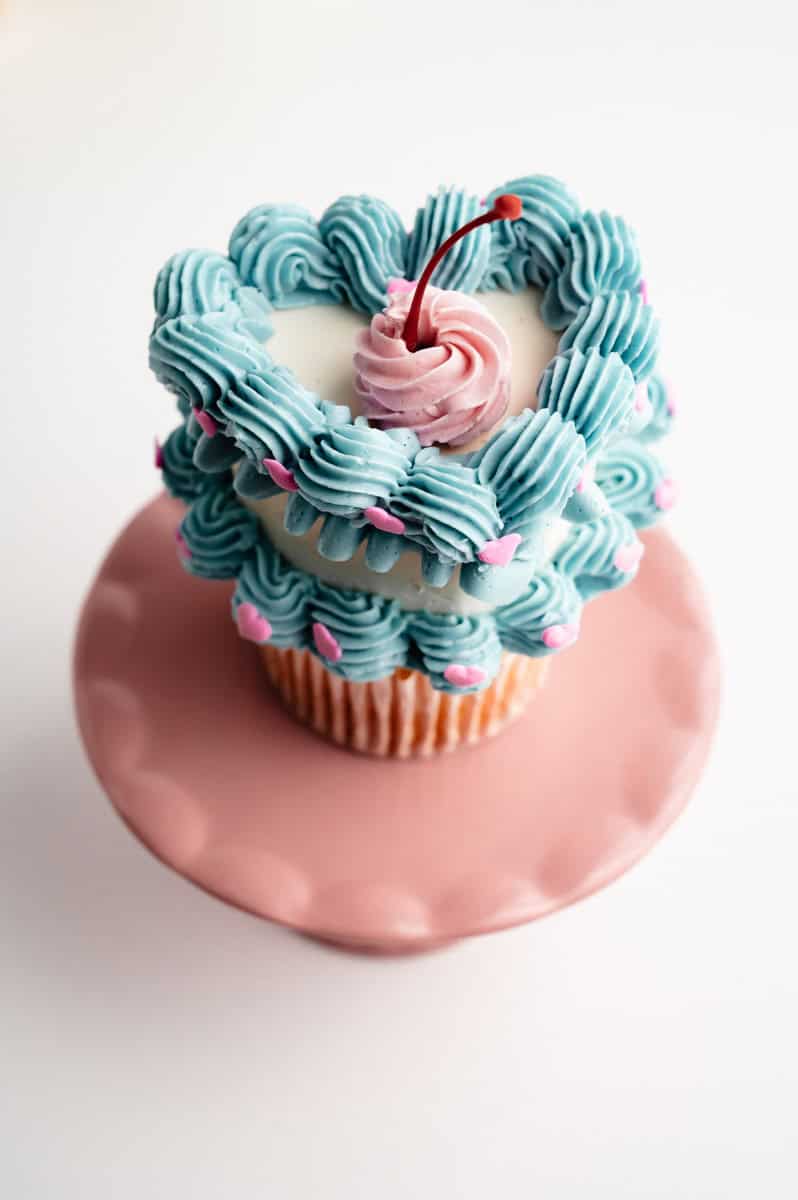 A vintage-style cupcake with a white heart buttercream decoate heart. The outside of the heart cupcake is a blue ribbon and blue scalloped edge details, with pink heart sprinkles in between each buttercream shell. On top is a pink buttercream swirl with a red Maraschino cherry stem.