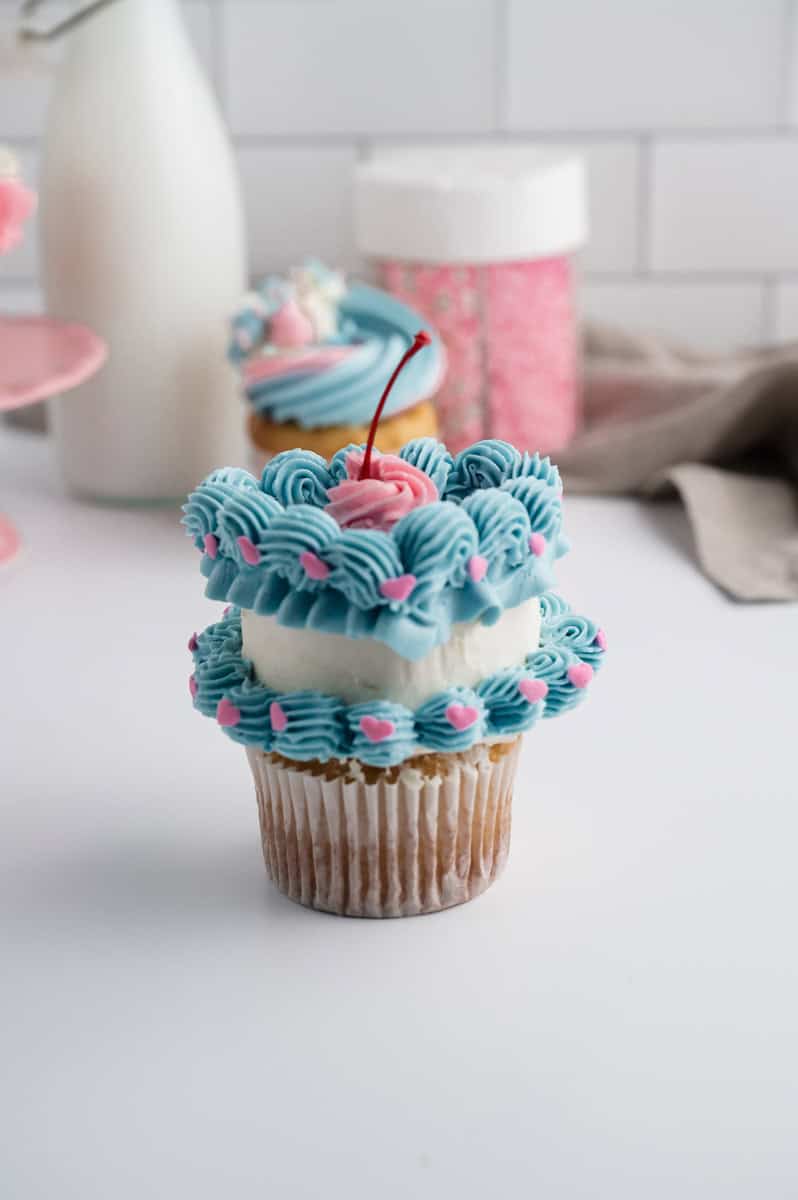 Heart cupcake in front of with pink sprinkles and light beige dish towel. There is also a vanilla cupcake with swirls of blue, pink, and white buttercream .