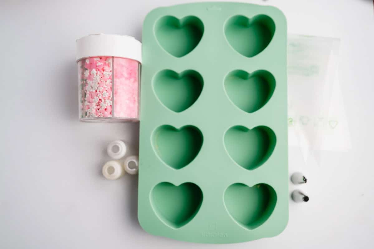 Collection of supplies and ingredients: silicone heart mold, piping couplers, cake decorating piping bags, sprinkles, and piping tips.