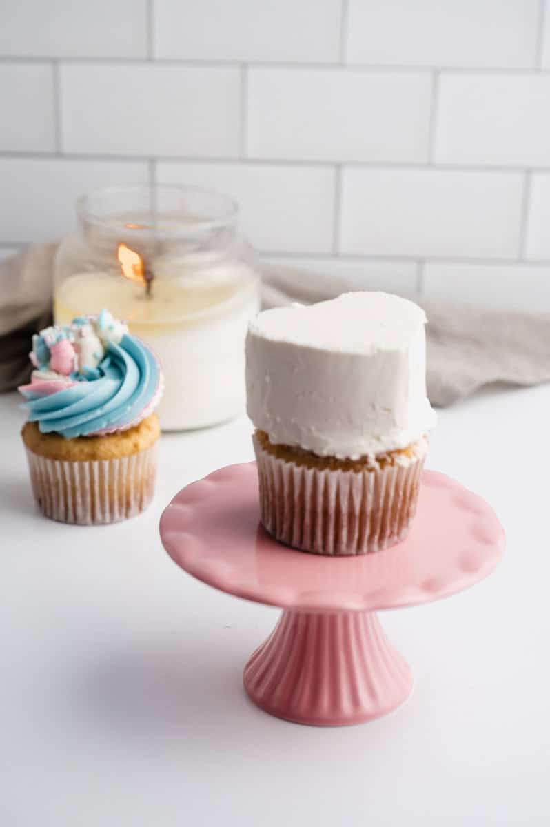 White buttercream heart cupcake is on top of a pink cupcake stand. No decorating or buttercream has been added to the white base. Vanilla cupcake, white candle, and beige dish towel are in background.
