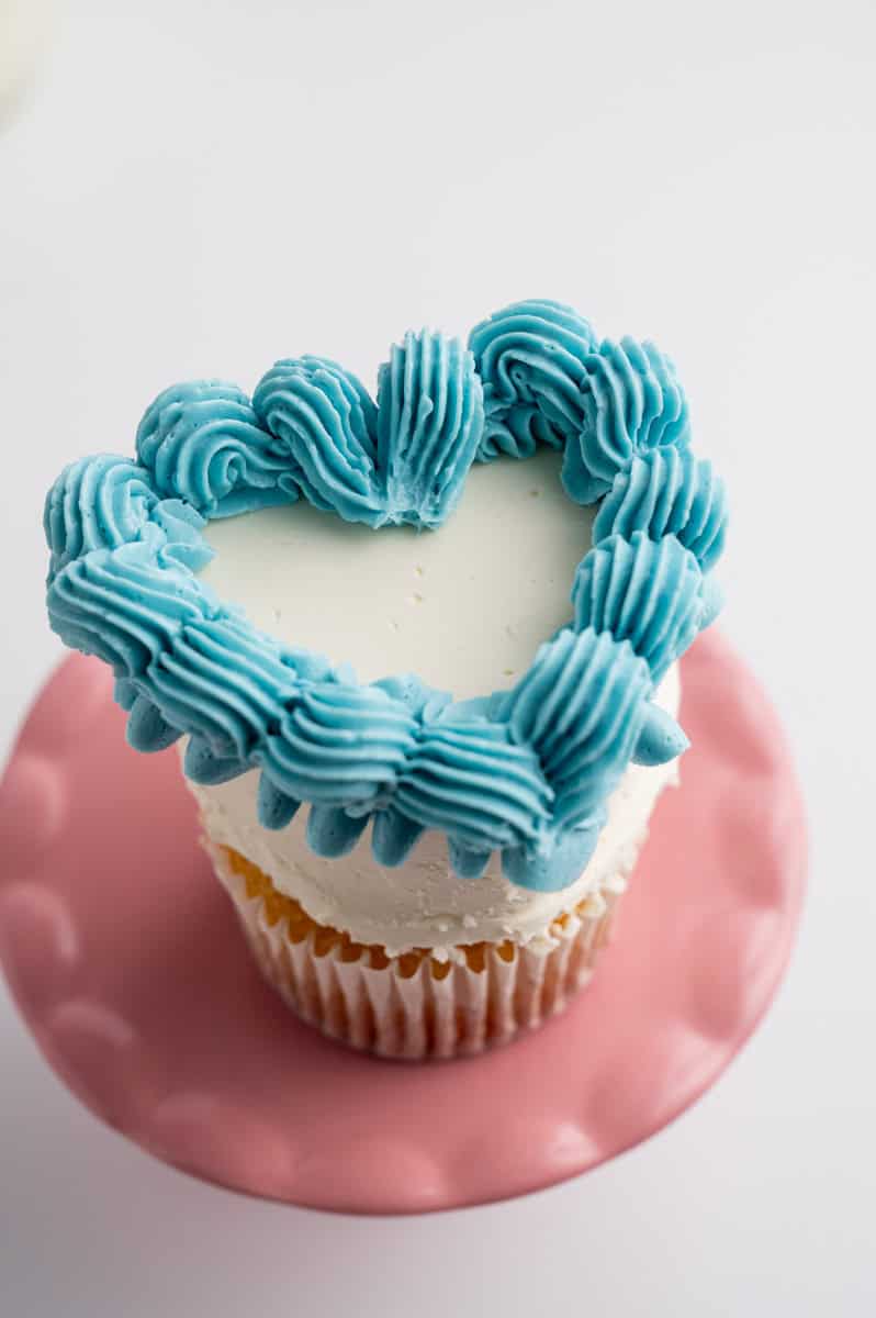 The white cupcake features a blue shell border added on top of the ribbon. The outer border of the cupcake is adorned with a ribbon, complemented by a matching blue shell border along the top.