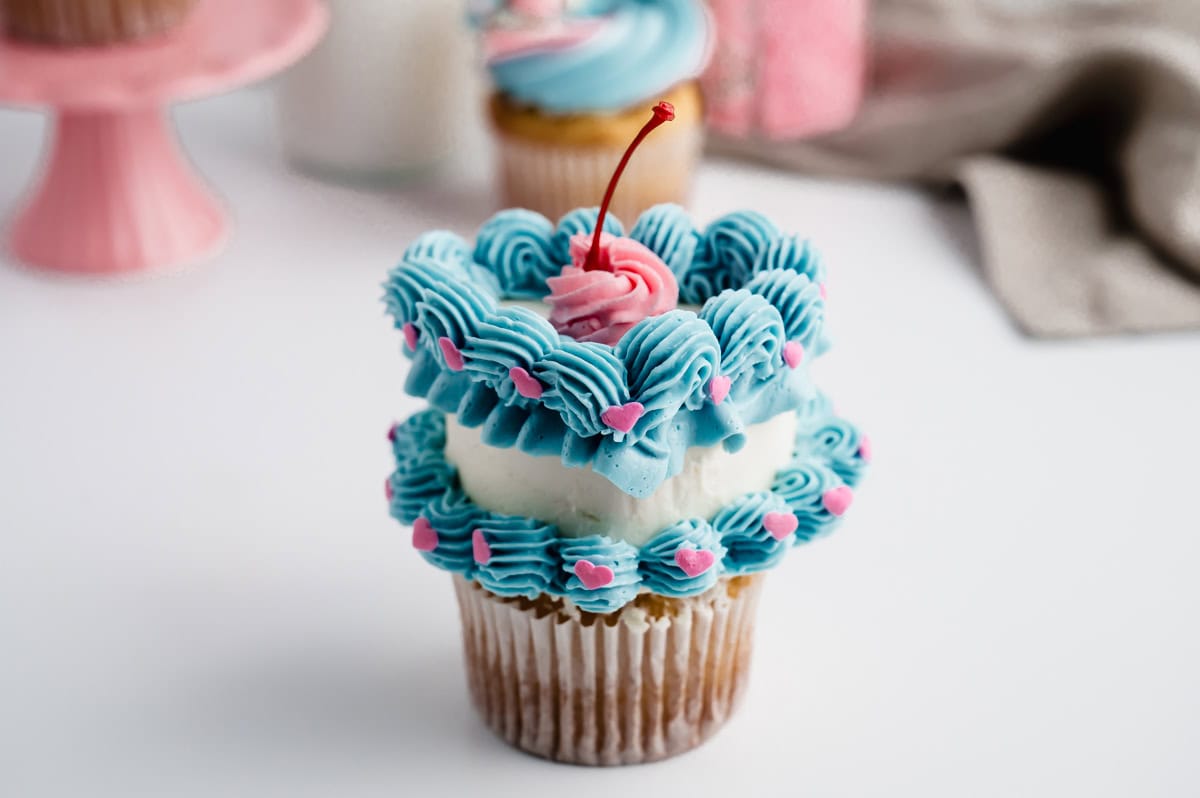 Front profile of the cupcake with white buttercream heart adorned with lower and upper rows of blue scalloped borders. The upper border has a blue ribbon base. A swirl of pink buttercream is in the center of the cupcake with a red maraschino cherry stem.