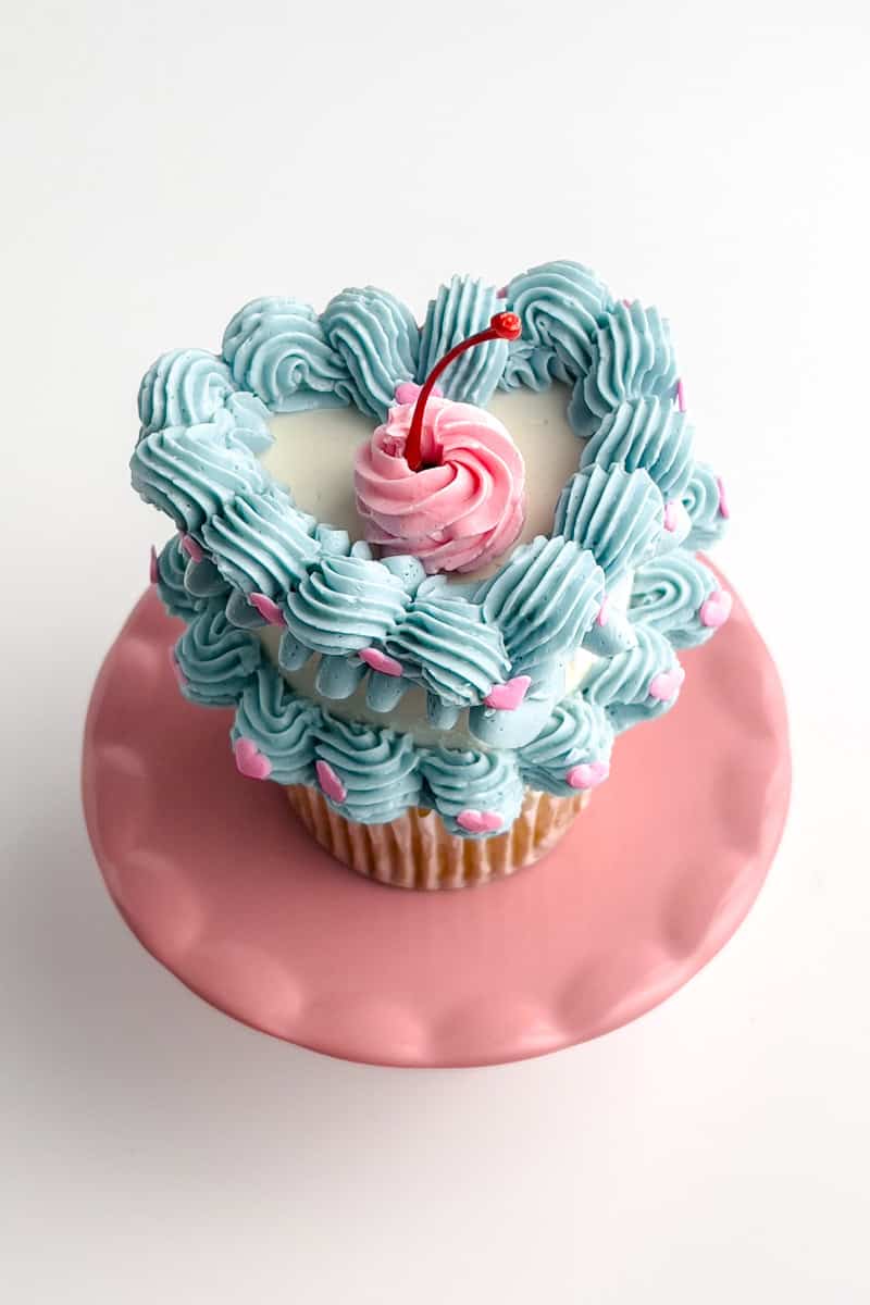 Front profile of the cupcake with white buttercream heart adorned with lower and upper rows of blue scalloped borders. The upper border has a blue ribbon base. A swirl of pink buttercream is in the center of the cupcake with a red maraschino cherry stem.