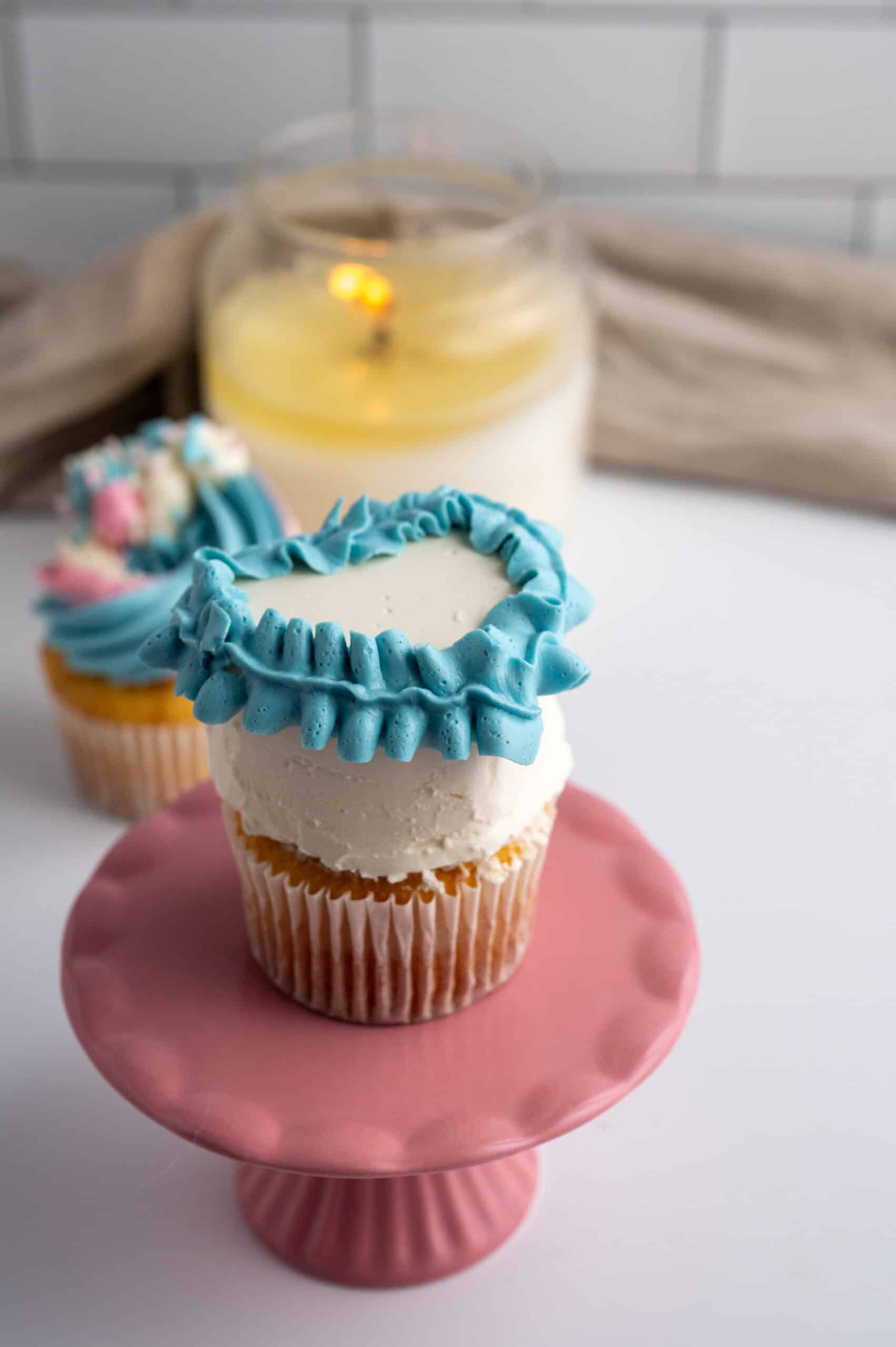 White cupcake has blue ribbon added to the top border of the cupcake. Cupcake and candle are in the background.