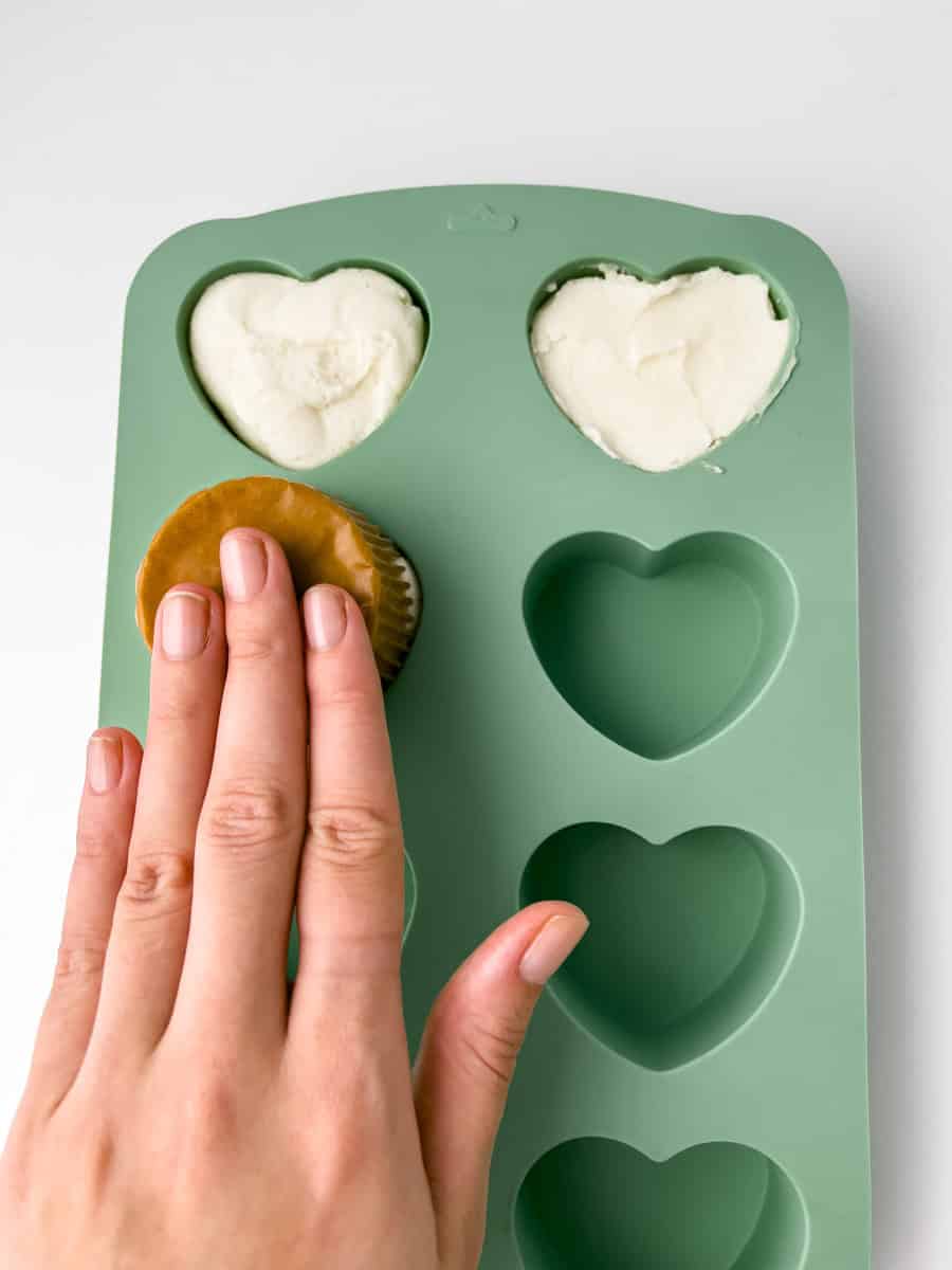 The palm of a hand is pushing down onto the bottom of a cupcake. The cupcake is pressed into a buttercream filled heart silicone mold.
