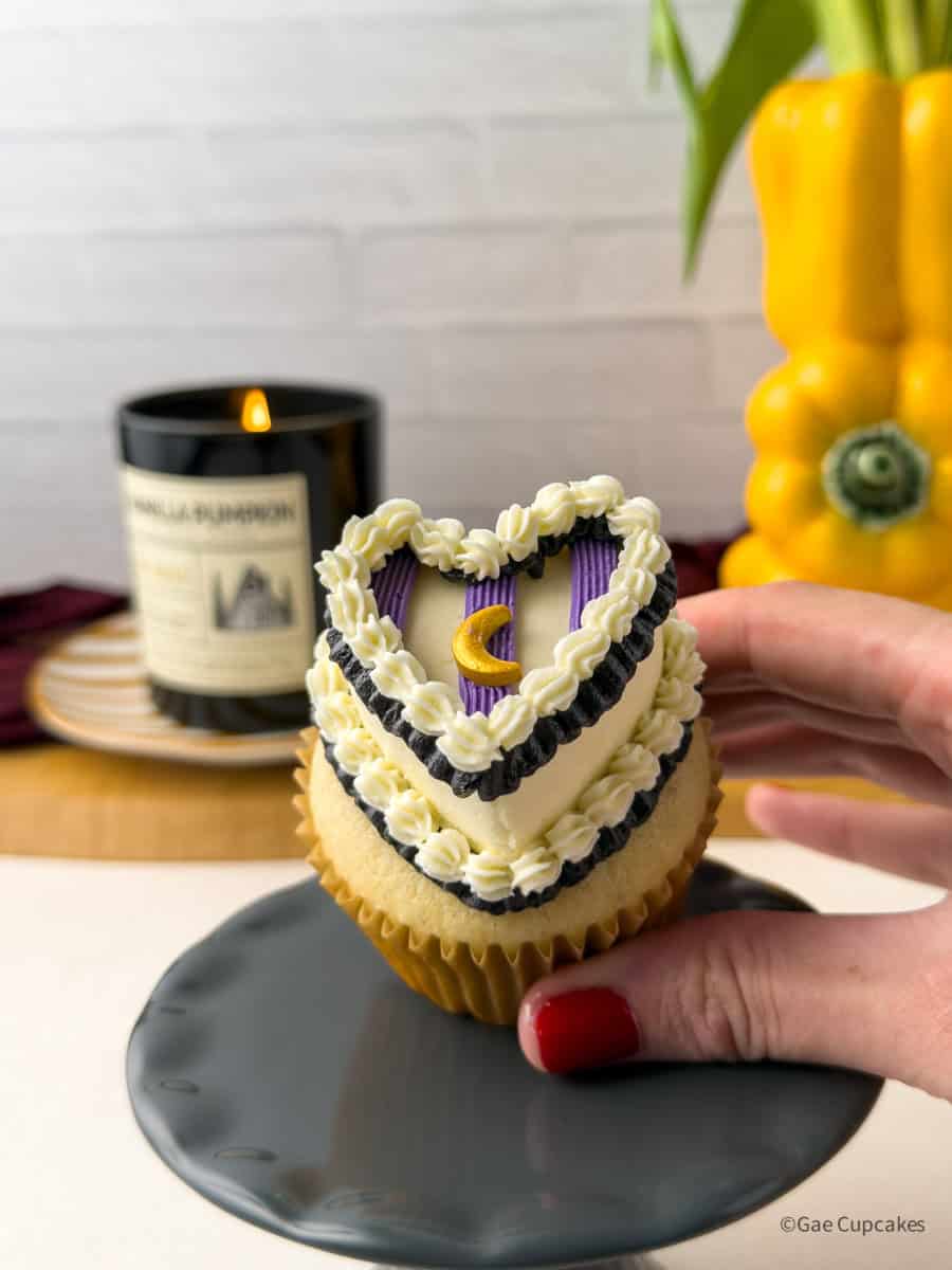 Witchy heart cupcake with vintage buttercream piping in white, black, and purple, topped with a gold moon sprinkle, held against a moody styled background with a lit candle.