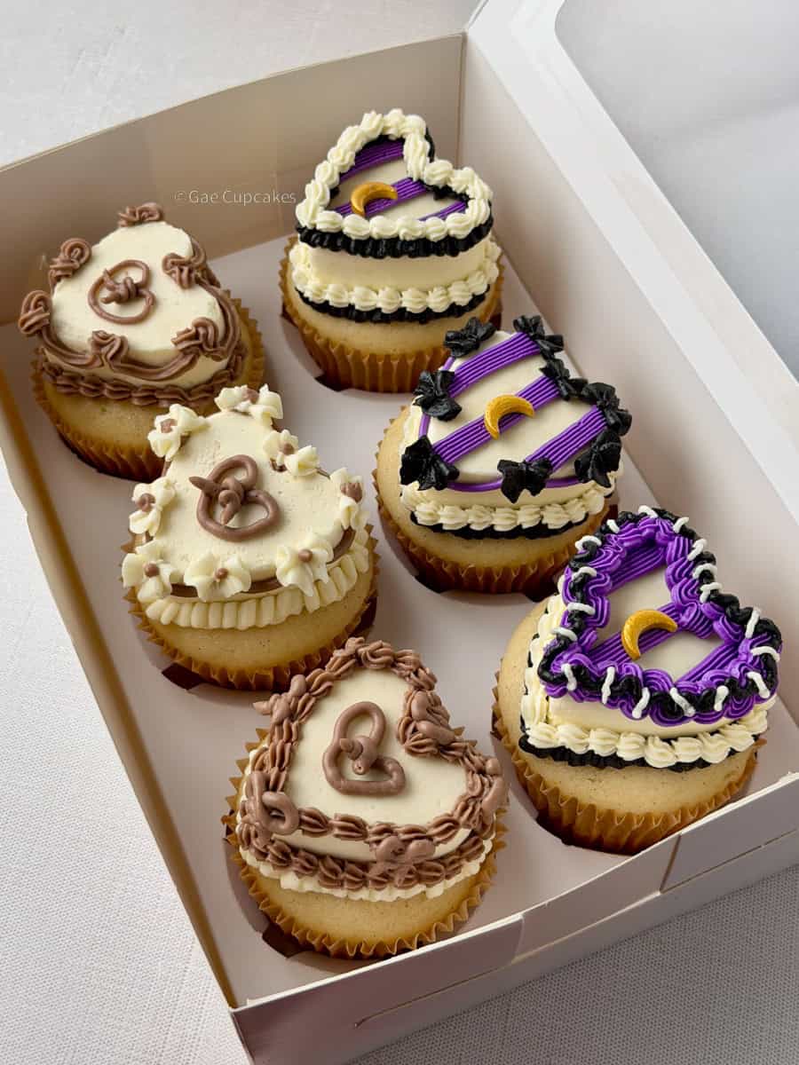 Gae Cupcakes decorated cupcakes in a white bakery box featuring witchy Halloween designs in purple and black with crescent moons, and vintage-style pretzel cupcakes in cream and chocolate brown frosting.