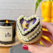 Hand holding a vanilla cupcake in a brown liner topped with a white buttercream heart and a black ruffled border at its base, with three purple stripes piped across the surface in equal thirds. A black ruffled border and white shell border are piped across the top of the purple stripes, with a gold moon sprinkle placed on the center stripe. A black candle and a yellow bell pepper used as a vase are visible in the background. Complete image of witchy heart cupcake simple style.