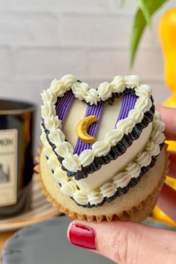 Hand holding a vanilla cupcake in a brown liner topped with a white buttercream heart and a black ruffled border at its base, with three purple stripes piped across the surface in equal thirds. A black ruffled border and white shell border are piped across the top of the purple stripes, with a gold moon sprinkle placed on the center stripe. A black candle and a yellow bell pepper used as a vase are visible in the background. Complete image of witchy heart cupcake simple style.