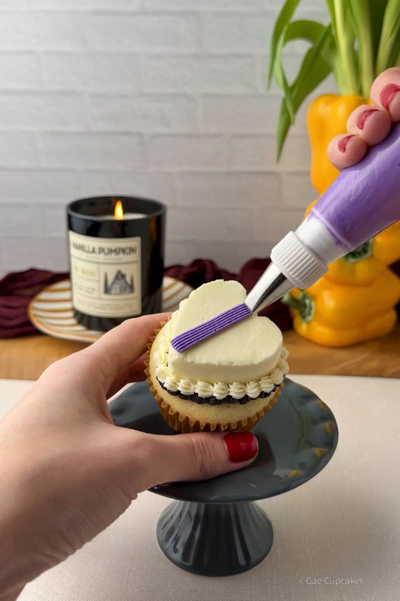 Step eight of witchy heart cupcake decorating tutorial. A vanilla cupcake in a brown liner topped with a white buttercream heart and a black ruffled border at its base, with a purple stripe piped down the center. A black candle and a yellow bell pepper used as a vase are visible in the background.