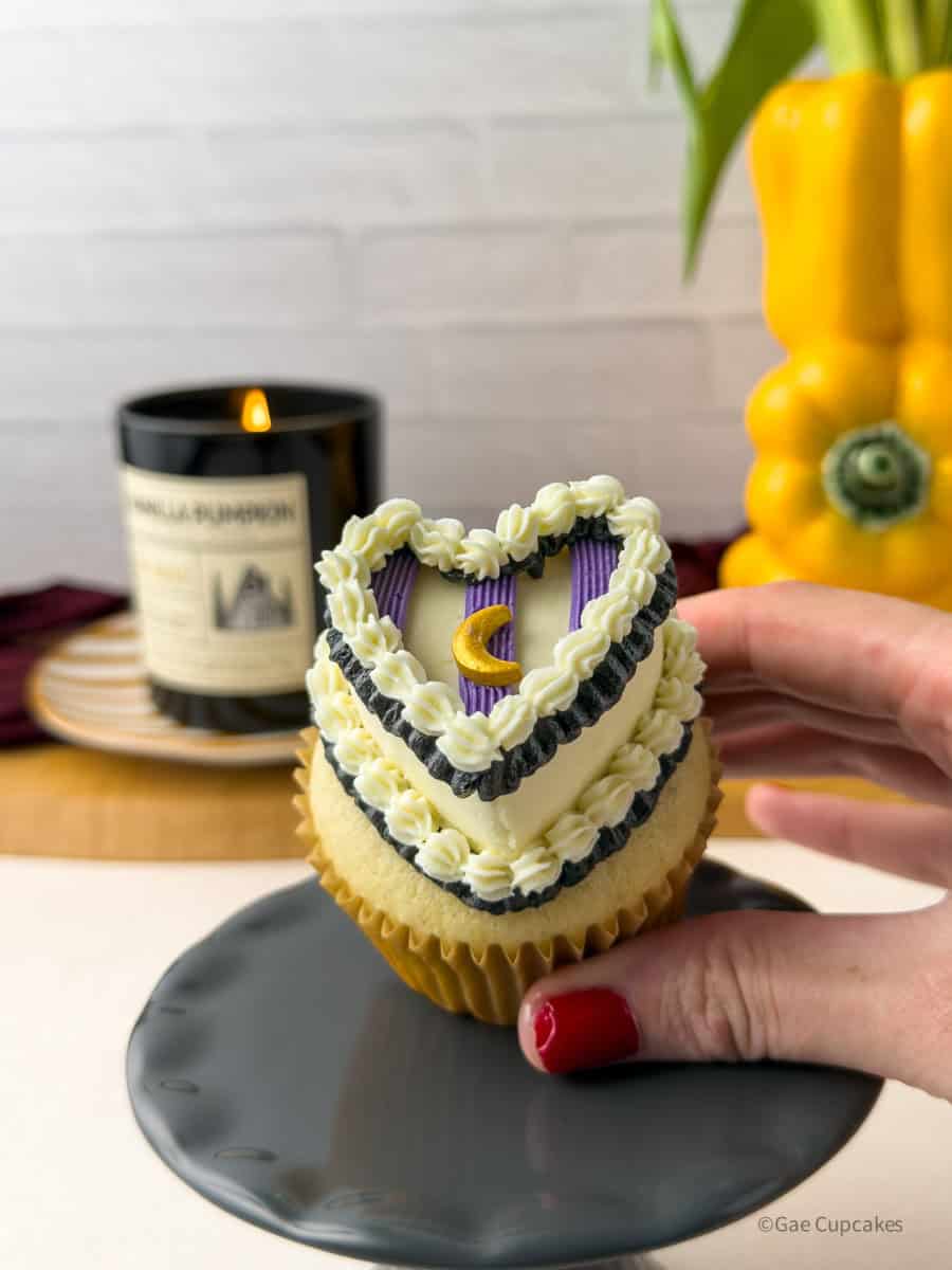Step Eleven of the witchy heart cupcake tutorial. A vanilla cupcake in a brown liner topped with a white buttercream heart and a black ruffled border at its base, with three purple stripes piped across the surface in equal thirds. A black ruffled border and white shell border are piped across the top of the purple stripes, with a gold moon sprinkle placed on the center stripe. A black candle and a yellow bell pepper used as a vase are visible in the background.