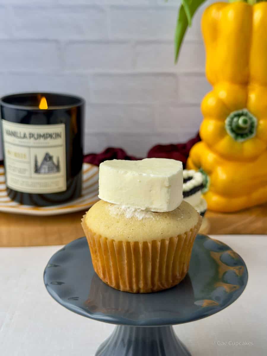 Step Five. A vanilla cupcake in a brown liner topped with a white buttercream heart. A black candle and a yellow bell pepper used as a vase are visible in the background.