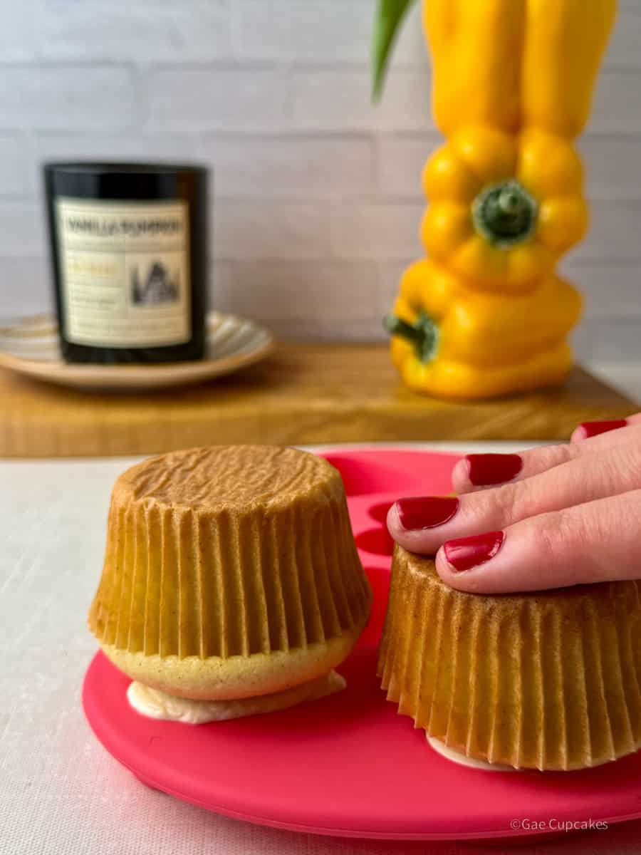 Step Four.Hands pressing cupcakes top-down into a pink silicone heart-shaped mold filled with white buttercream. A black candle and a yellow bell pepper used as a vase are visible in the background.