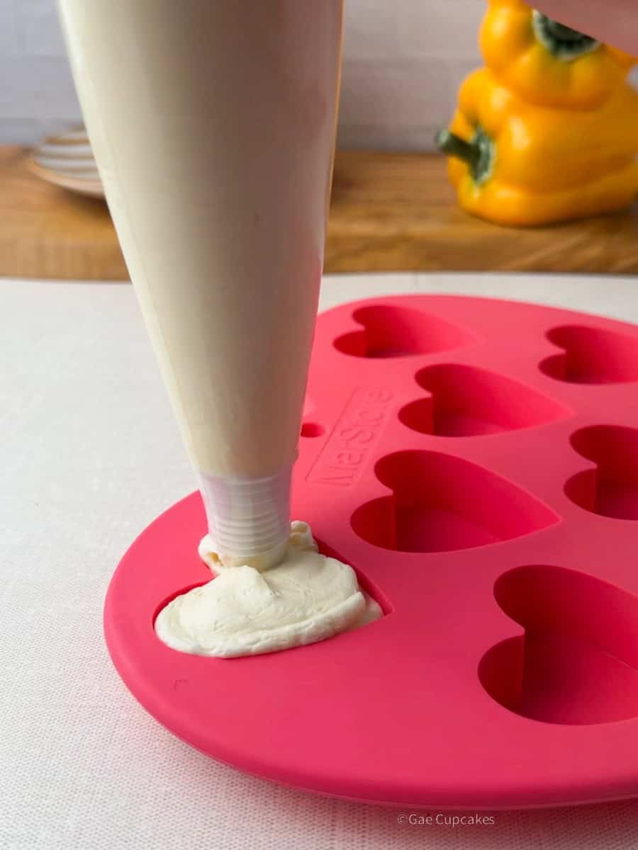 Step One of witchy heart cupcake. Pink silicone heart-shaped mold being filled with white buttercream from a white piping bag. A black candle and a yellow bell pepper used as a vase are visible in the background.