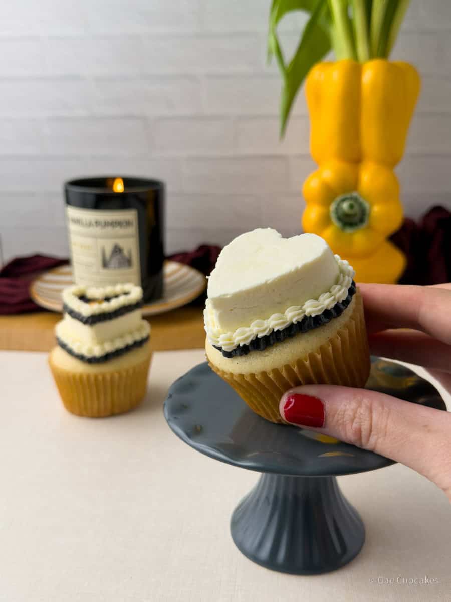 Step seven of witchy heart cupcake decorating tutorial. A vanilla cupcake in a brown liner topped with a white buttercream heart and a black ruffled border at its base. On top of the black ruffle is a white shell border. A black candle and a yellow bell pepper used as a vase are visible in the background.