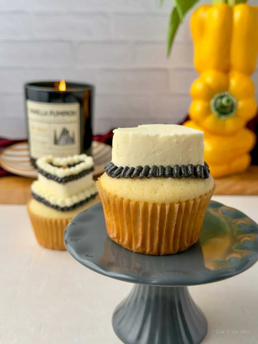 Step Six of witchy heart cupcake decorating tutorial. A vanilla cupcake in a brown liner topped with a white buttercream heart and a black ruffled border at its base. A black candle and a yellow bell pepper used as a vase are visible in the background.