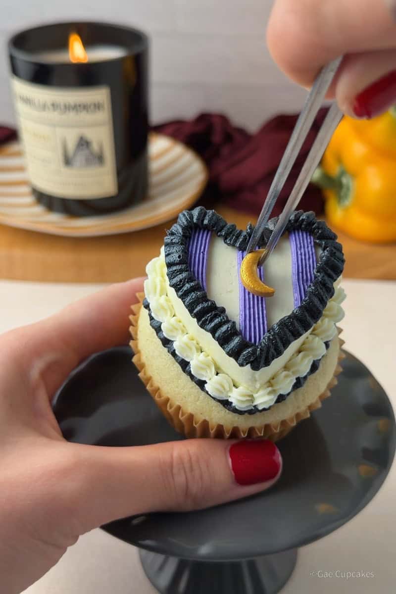 Step 10 of the witchy heart cupcake decorating tutorial. A vanilla cupcake in a brown liner topped with a white buttercream heart and a black ruffled border at its base, with three purple stripes piped across the surface in equal thirds and a gold moon sprinkle placed on the center stripe. A black candle and a yellow bell pepper used as a vase are visible in the background.